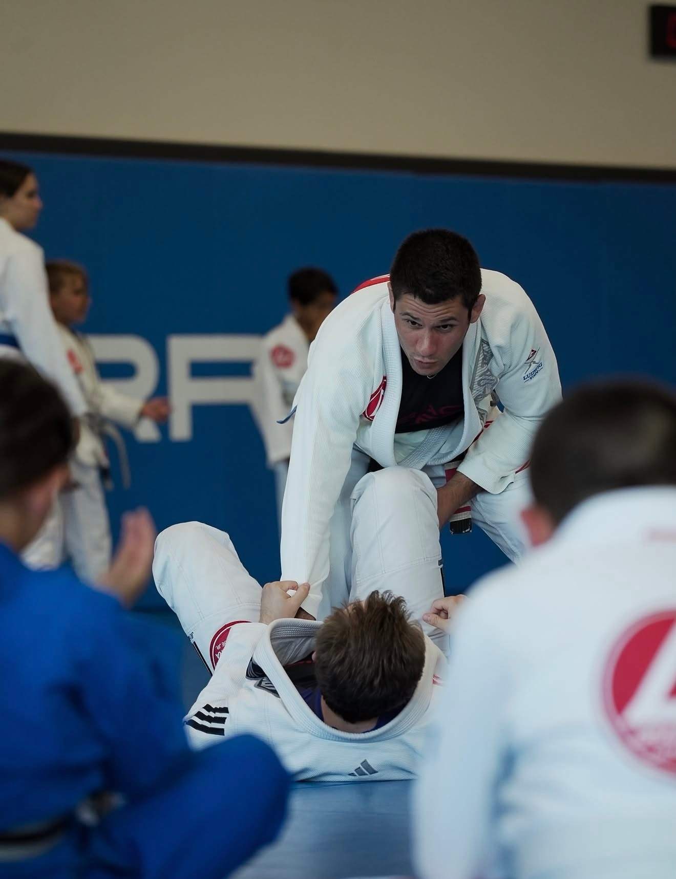 An instructor demonstrates a technique on a partner lying on the mat while students watch