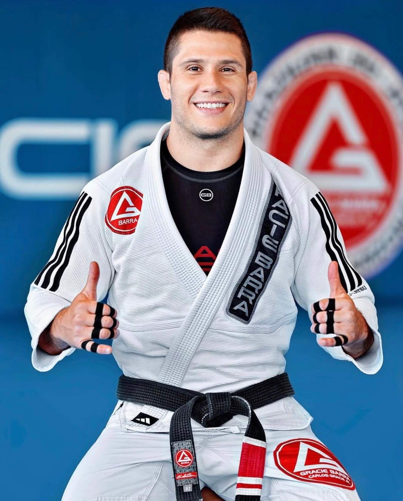 Head Professor Lucas Rubbo, in a white Gracie Barra gi and black belt, smiles and gives two thumbs up in front of the Gracie Barra logo