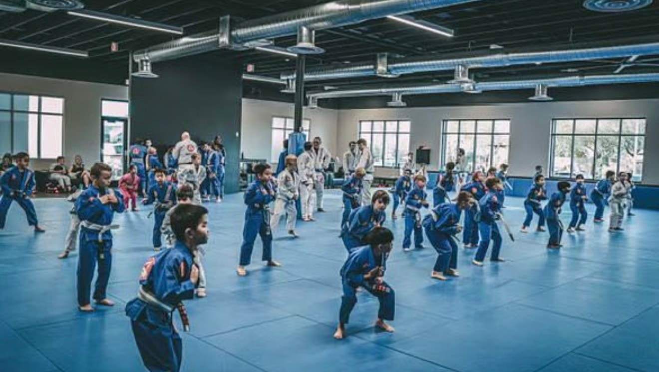 A large kids class in blue and white gis warms up across the mats, coaches and parents in the background
