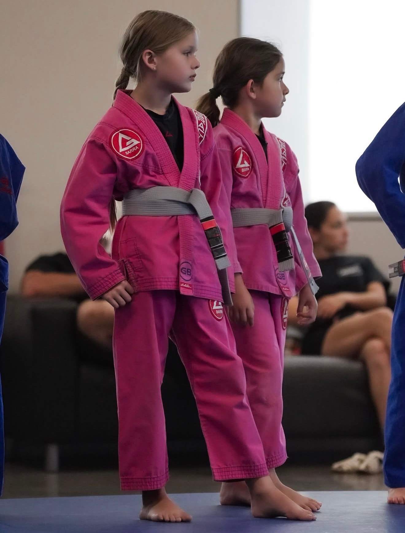 Two young girls in pink gis and gray belts stand in line at the start of class, parents seated behind them
