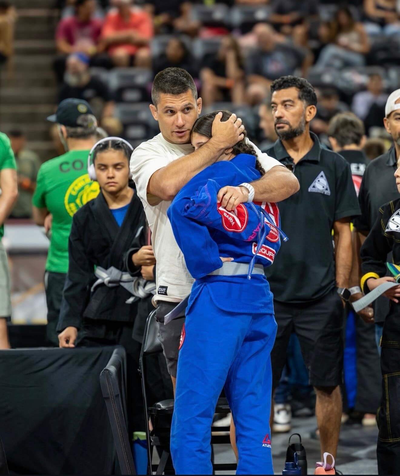 A coach hugs a young competitor in a blue Gracie Barra gi at a jiu-jitsu tournament